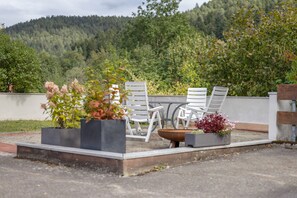 Un área de estar al aire libre con sillas y mesas blancas, rodeada de macetas con flores, con un paisaje de colinas boscosas de fondo.
