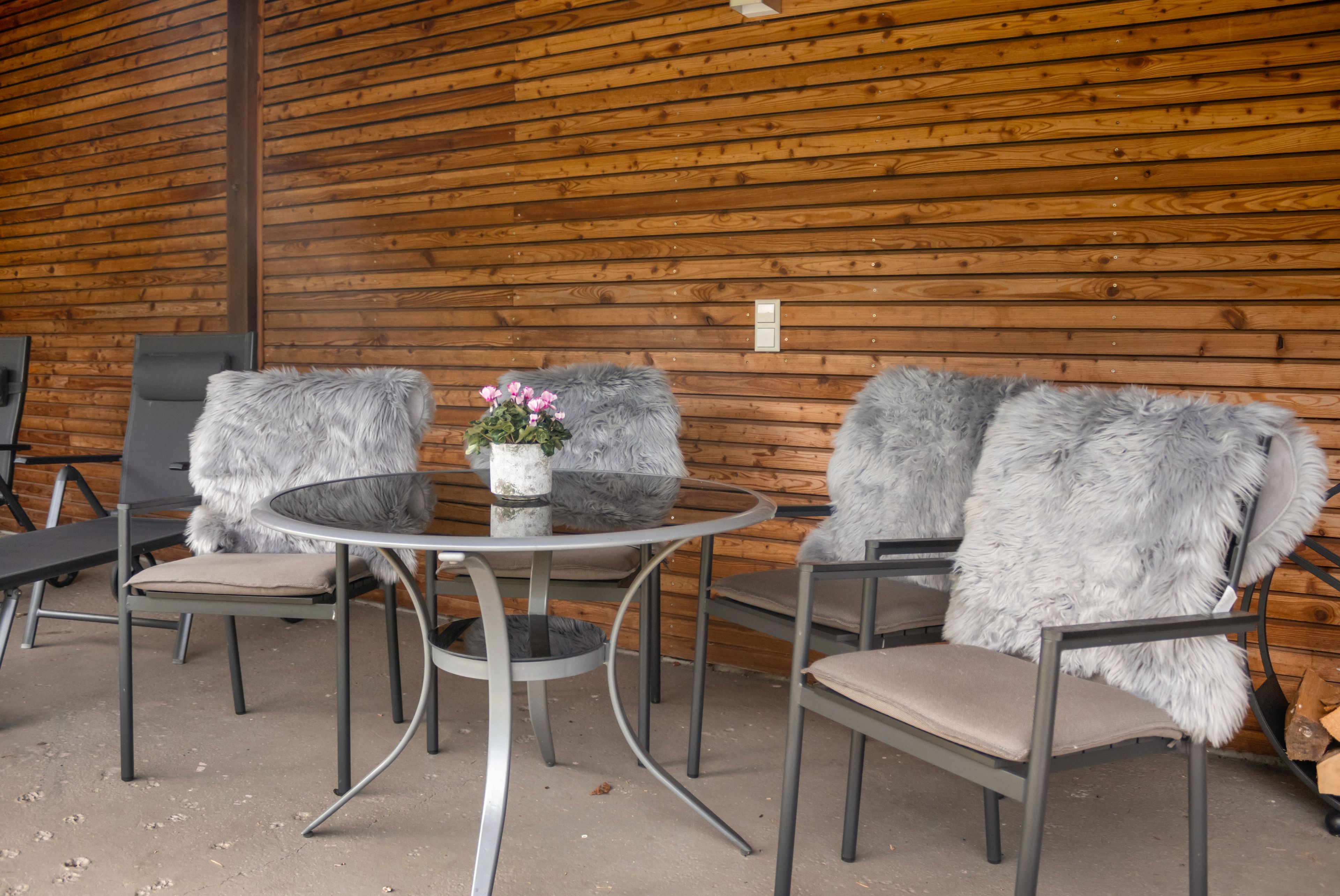 A round metal table with two chairs, each with a fur throw. A small potted plant with pink flowers is on the table.
