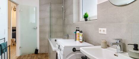 A modern bathroom with a glass shower enclosure, a washing machine, a sink, and a mirror.