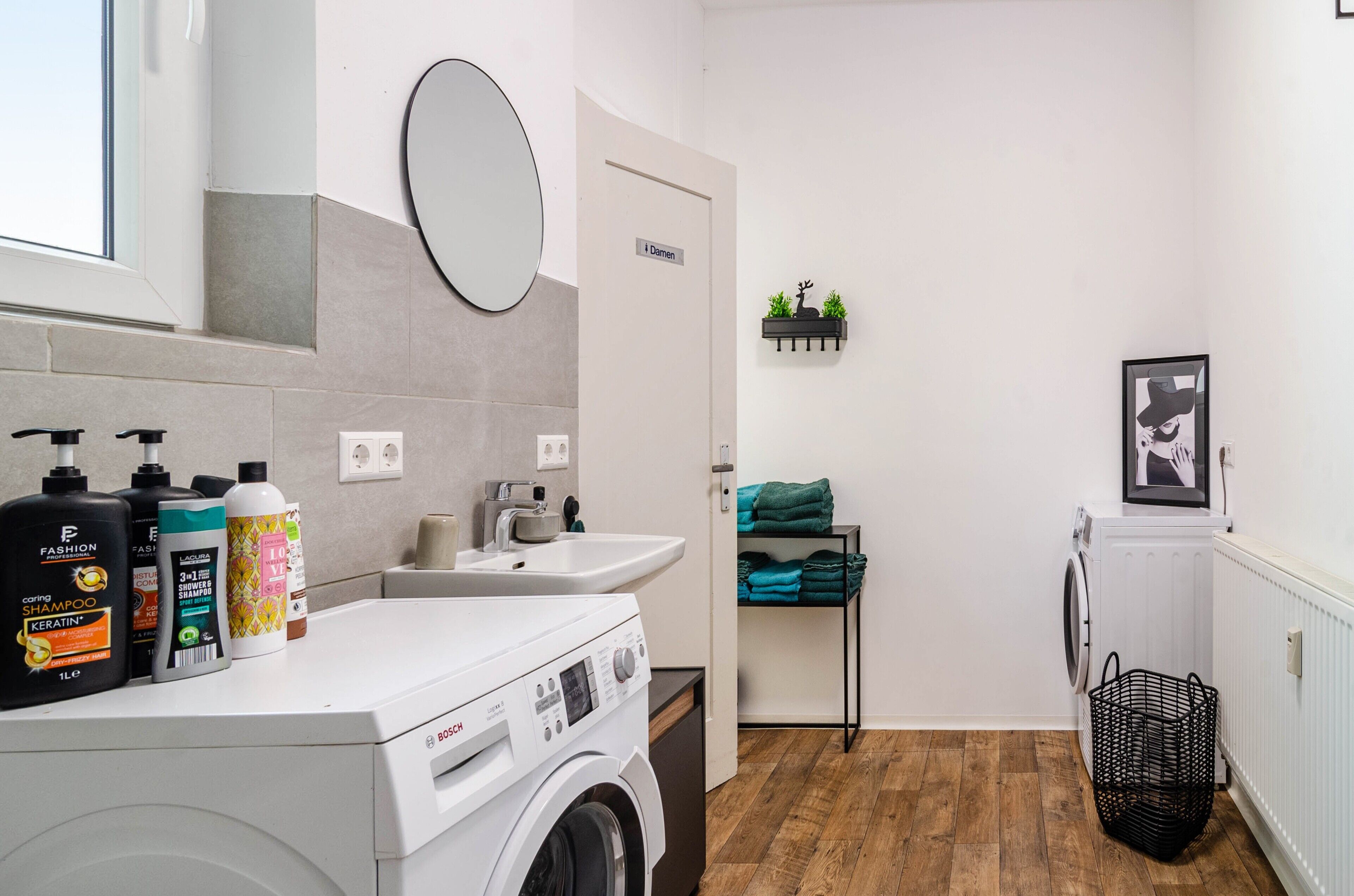 A modern laundry room with a Bosch washing machine, a sink, a mirror, and a door labeled 'Damen.'
