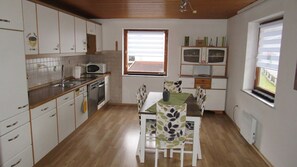 Een kleine keuken met een eethoek, houten keukenkastjes, een magnetron en een raam met zonwering.