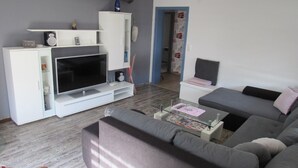 A living room with a flat-screen TV, a grey sofa, a white entertainment unit, and a door with a red car decal. - Apartment 'Schopper' with Balcony and Wi-Fi (Halblech)