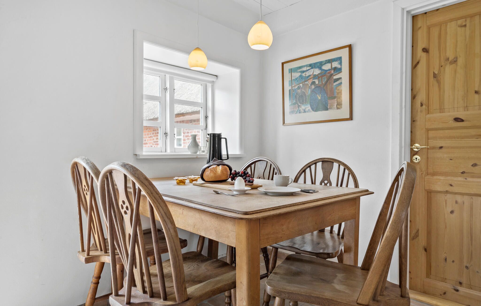 A dining area with a wooden table and chairs, a window with a view of a brick building, a painting on the wall, and a door with a wooden panel.