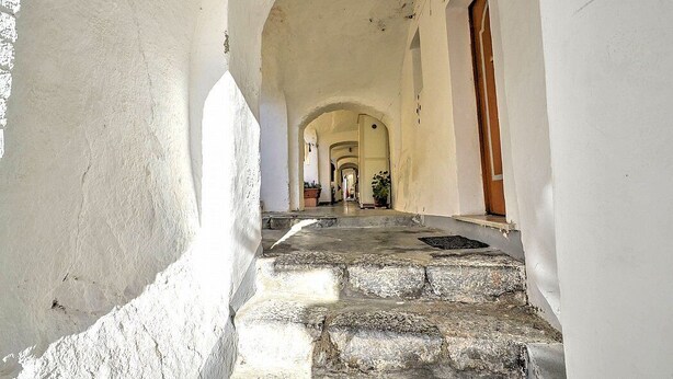 Un couloir avec des marches en pierre menant à une porte en arc de cercle, flanquée de murs blancs et d’une porte en bois.
