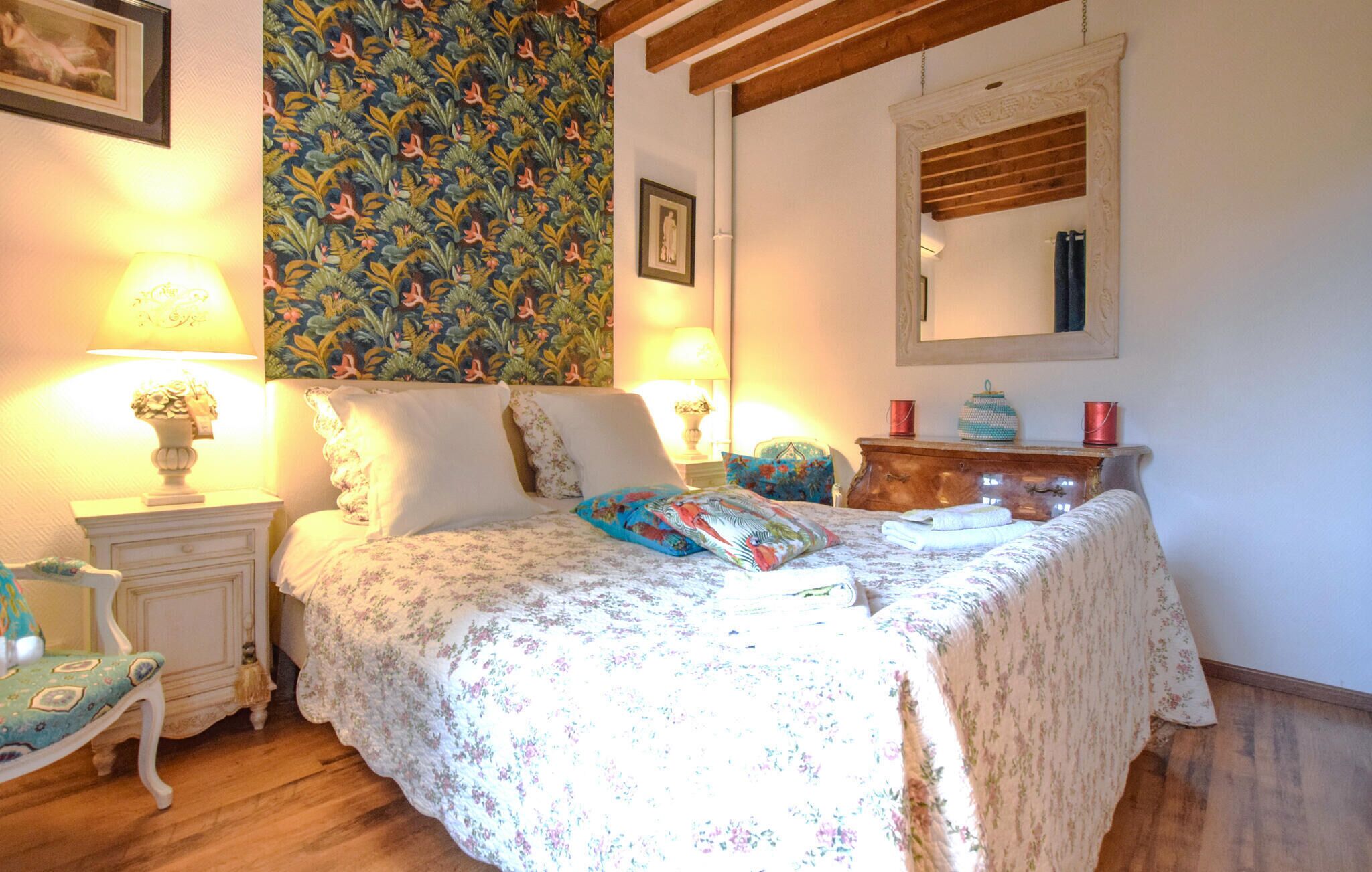 A bedroom with a floral bedspread, a wooden dresser, two bedside lamps, a chair, and a patterned wallpaper.