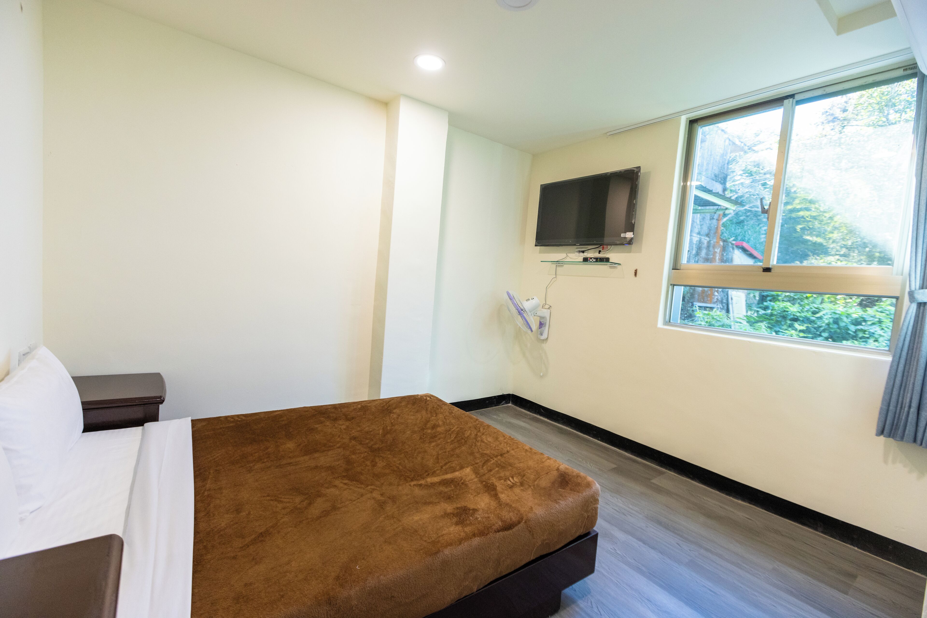 A hotel room with a bed, a TV mounted on the wall, and a window with a view of greenery.