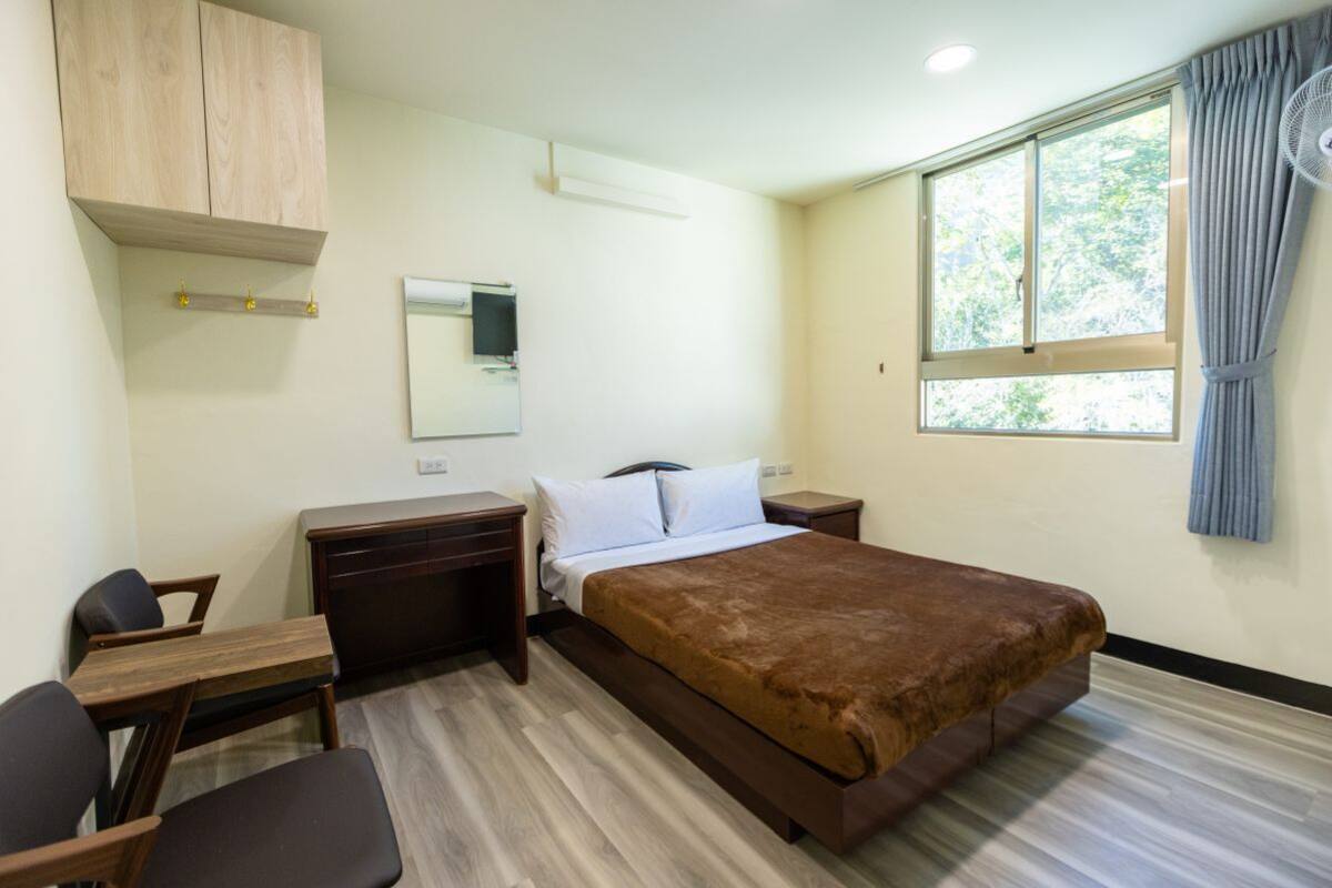 Habitación de hotel con cama, escritorio, silla, armario, espejo y ventana con cortinas.