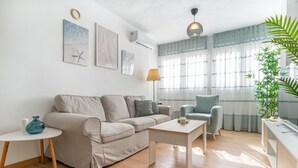 Ein modernes Wohnzimmer mit einem beigen Sofa, einem weißen Couchtisch, einem grauen Sessel und einem kleinen Beistelltisch mit einer blauen Vase. An der Wand hängen drei gerahmte Kunstwerke, eine Stehlampe und ein großes Fenster mit durchsichtigen Vorhängen.