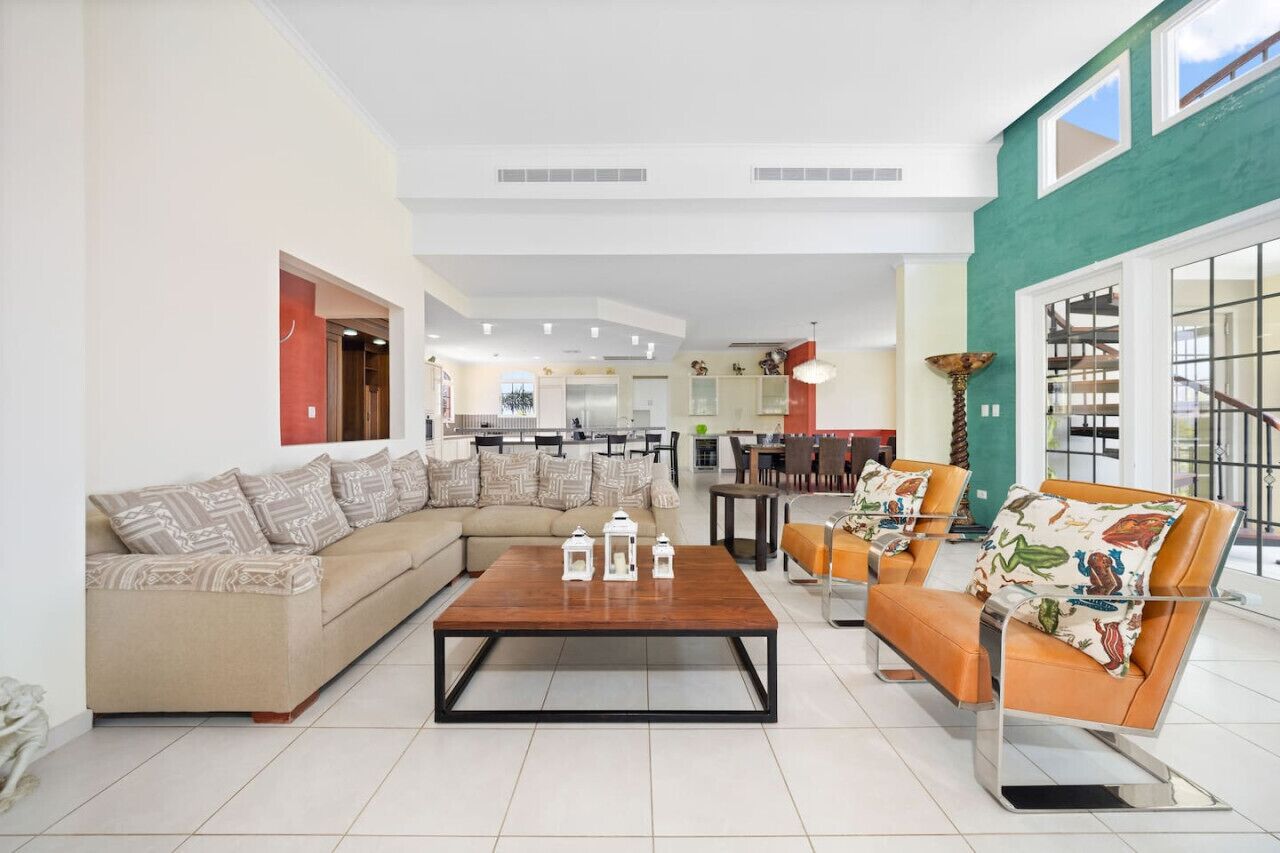 A spacious living area with a sofa, coffee table, and chairs, a kitchen in the background, and large windows.