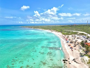 Una playa costera con aguas turquesas cristalinas, una orilla de arena y una hilera de estructuras con techo de paja a lo largo de la playa.