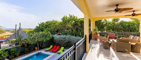 A poolside area with a view of the ocean, lounge chairs, and a covered patio with wicker furniture.