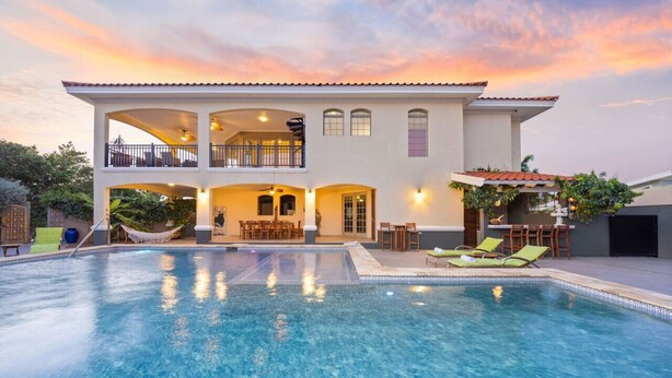 Una casa de dos pisos con piscina, mobiliario exterior y un balcón.