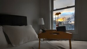 Ein Hotelzimmer mit einem Bett, einem Nachttisch aus Holz, einer Lampe und einem Fenster mit Blick auf Häuser und einen Sonnenuntergang.