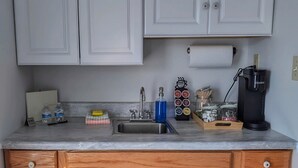 Eine Küche mit weißen Schränken, einem Spülbecken und Holzunterschränken. Auf der Arbeitsplatte befindet sich eine Kaffeemaschine, eine Flasche Spülmittel und ein Zuckerbehälter.