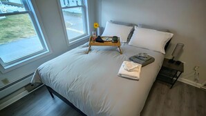 A bedroom with a large bed, a bedside table with a lamp, and a window with a view of the outside. - The Mast at Soundview · Beach+Ocean+Sunrise (Old Lyme)