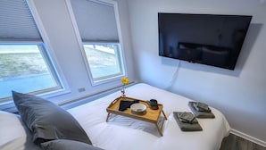 A bedroom with a bed, a bedside table with a tray, a TV mounted on the wall, and a window with blinds. - The Stern at Soundview · Beach+Ocean+Sunrise (Old Lyme)