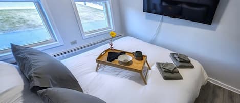 Un dormitorio con cama, una mesita de noche con bandeja, televisor montado en la pared y una ventana con persianas.