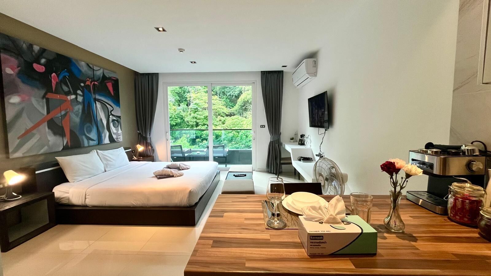 A modern hotel room with a large bed, a dining table, a TV, and a view of greenery outside.