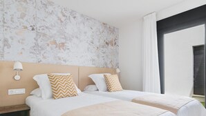 A hotel room with two beds, a nightstand, a wall-mounted lamp, and a window with curtains.