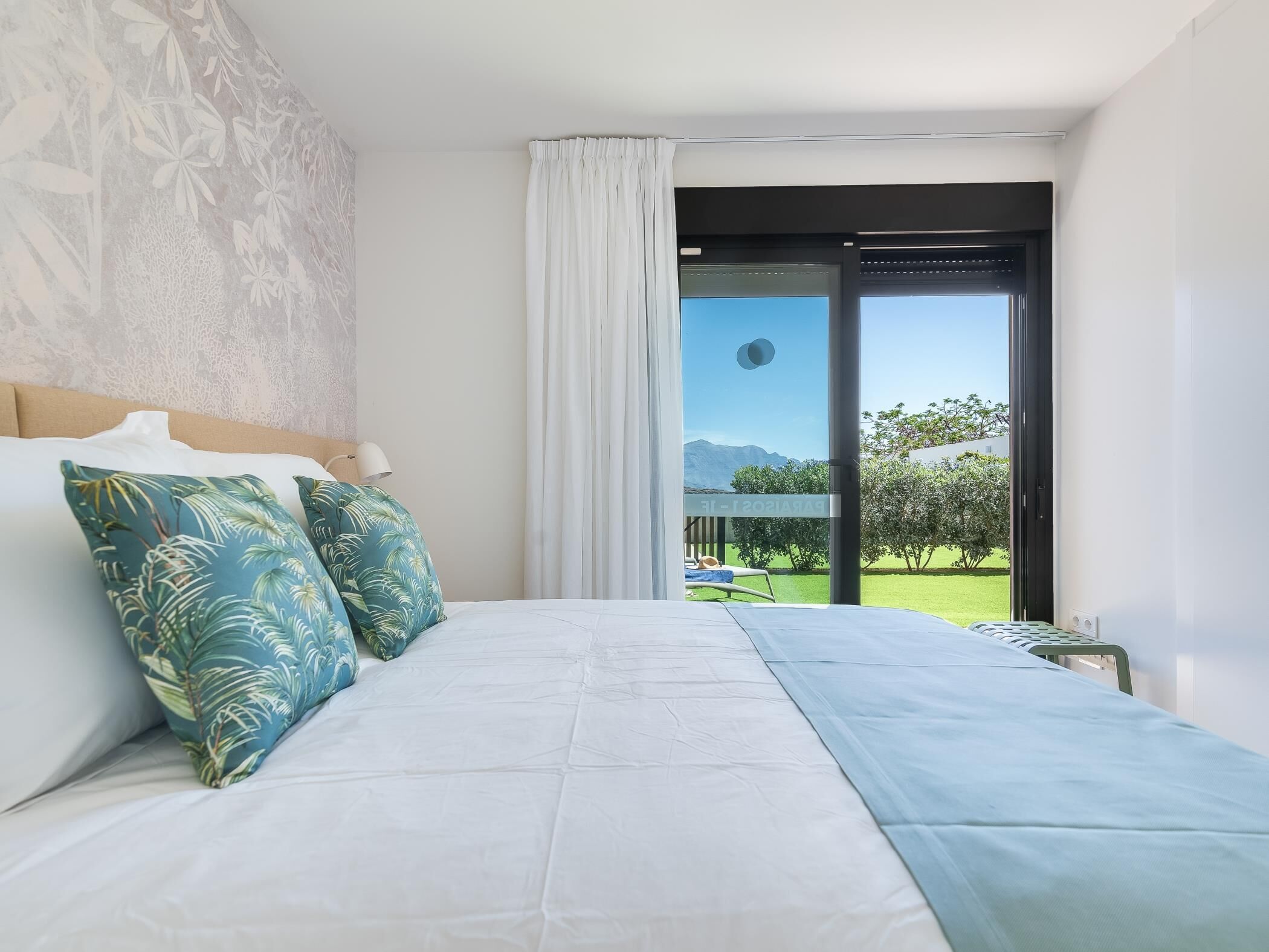Une chambre à coucher avec un lit, deux oreillers, une table de chevet et une vue sur un jardin, accessible par une porte coulissante.