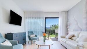A modern living room with a sofa, armchair, and a coffee table. A flat-screen TV is mounted on the wall, and there is a view of a garden through the window.