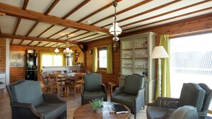 A cozy cabin interior with wooden beams, wicker furniture, a dining table, and a kitchen area with shelves. - Rendeux Valley View Retreat (Rendeux-Beffe)