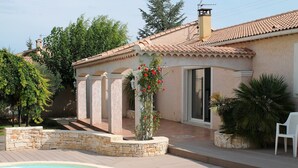 A pool area with a stone wall, a covered patio, and a house with a tiled roof. - Villa in Languedoc with Pool near Historic Cities (Rousson)