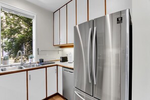 A modern kitchen with a stainless steel refrigerator, wooden cabinets, and a window with a view of greenery.