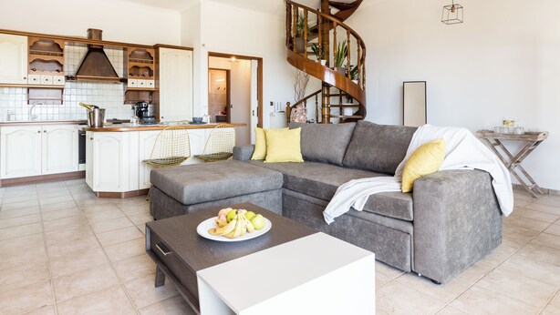 Una sala de estar moderna con un sofá seccional gris, una mesa de centro de madera con fruta, y una cocina al fondo que cuenta con gabinetes blancos y una escalera de madera.