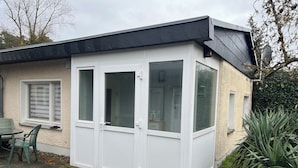 A white building with a black roof, a window with a white frame, and a door. There is a small table and chairs outside.