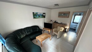 A living room with a black leather sofa, a wooden coffee table, and a beige armchair. There is a painting on the wall and a kitchenette in the background.