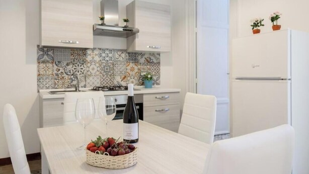 A modern kitchen with white cabinets, a tiled backsplash, and a dining table set with a bottle and fruit. - Housestation S (Palermo)