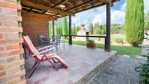 A covered patio area with a brick wall, a stone floor, and a view of a garden with a stone building and tall trees.