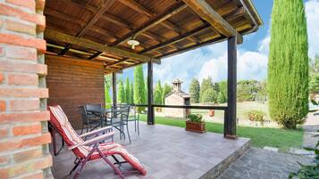 Ein überdachter Patio-Bereich mit Backsteinmauer, Steinboden und Blick auf einen Garten mit einem Steinhaus und hohen Bäumen.