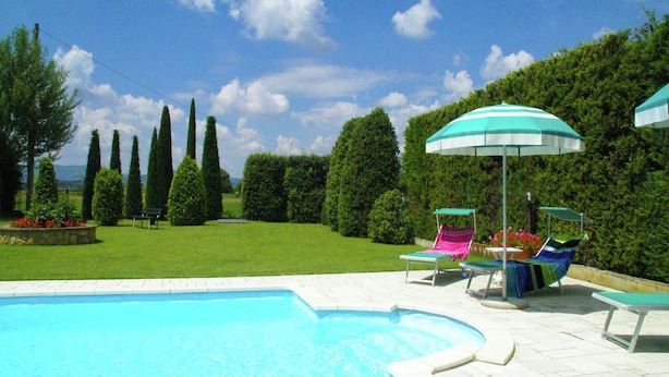 A swimming pool with lounge chairs and umbrellas, surrounded by neatly trimmed hedges and a clear sky.