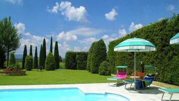 Ein Schwimmbecken mit Liegestühlen und Sonnenschirmen, umgeben von ordentlich geschnittenen Hecken und blauem Himmel.