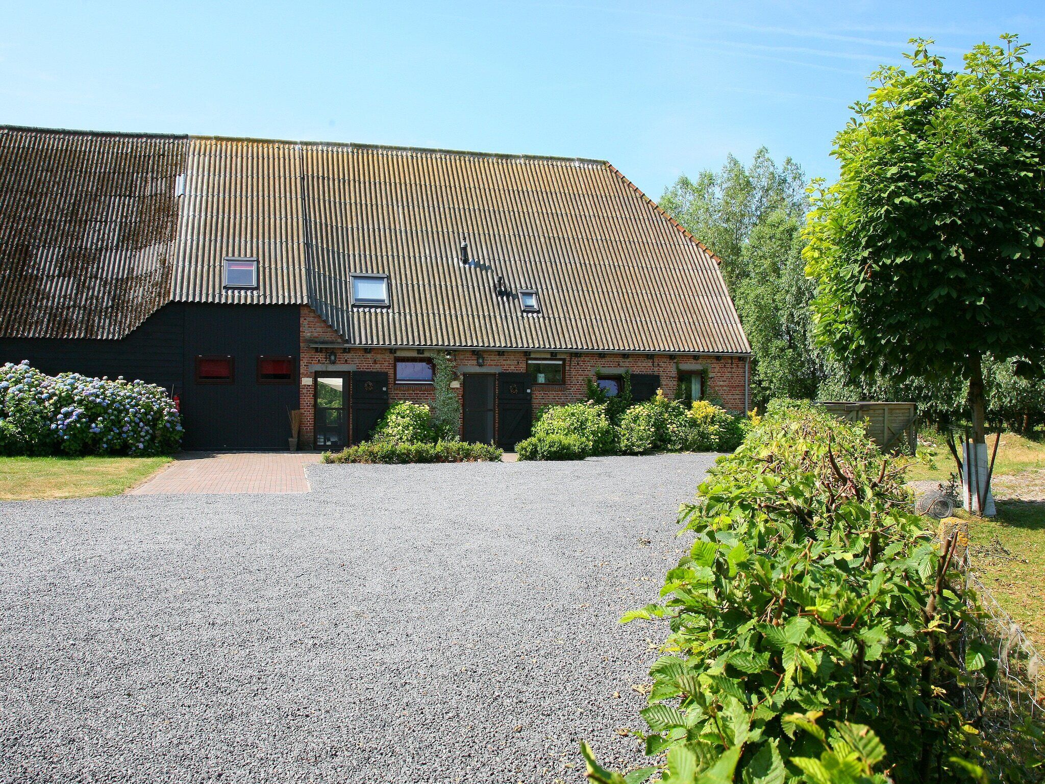 A building with a sloped roof, a gravel driveway, and a small garden area with bushes and a tree.