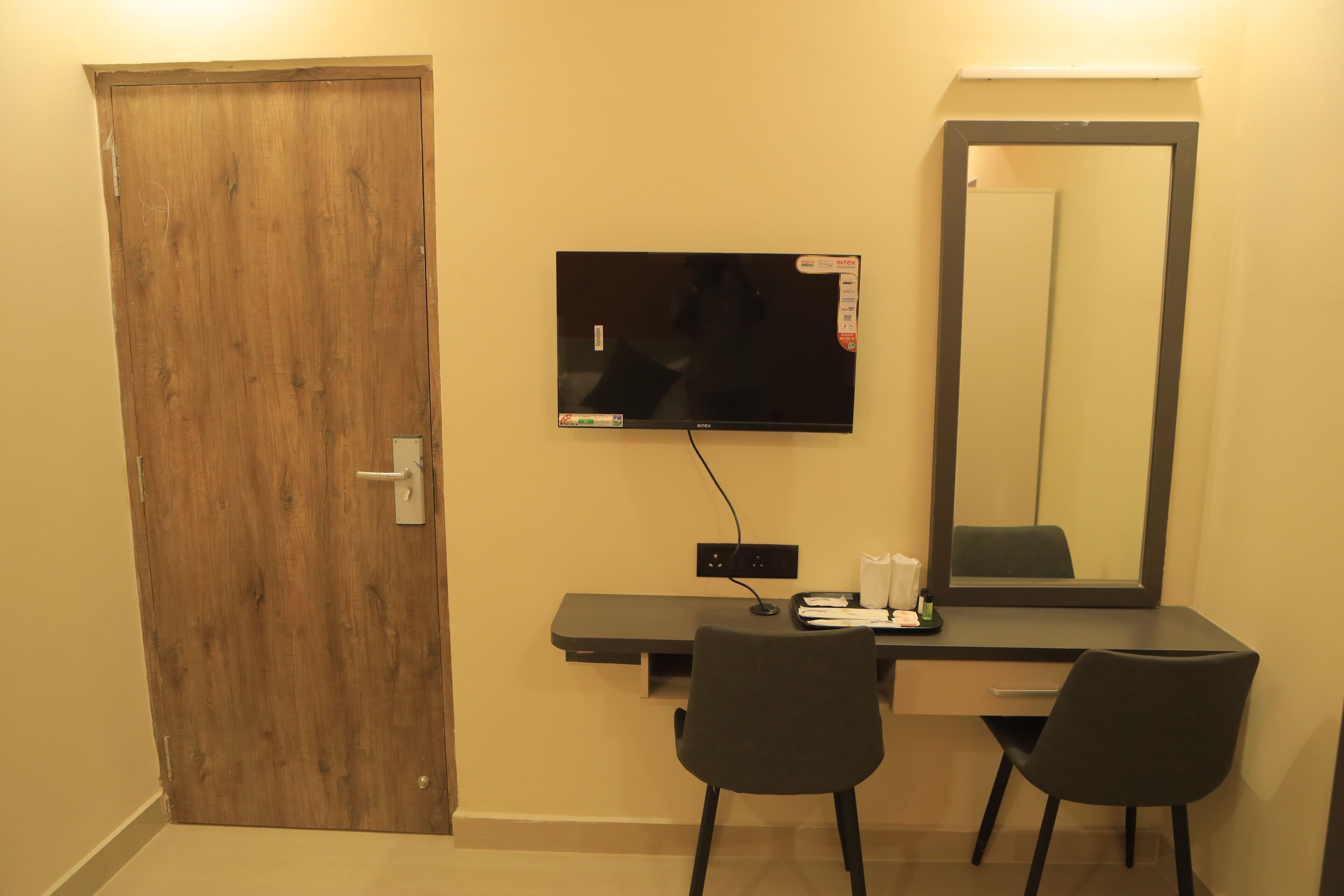 Ein Zimmer mit Holztür, einem flachbildfernseher an der Wand, einem Schreibtisch mit Stuhl und einem großen Spiegel.