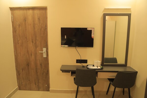 A room with a wooden door, a flat-screen TV mounted on the wall, a desk with a chair, and a large mirror. - Kansone Hotel And Banquets (Chennai)