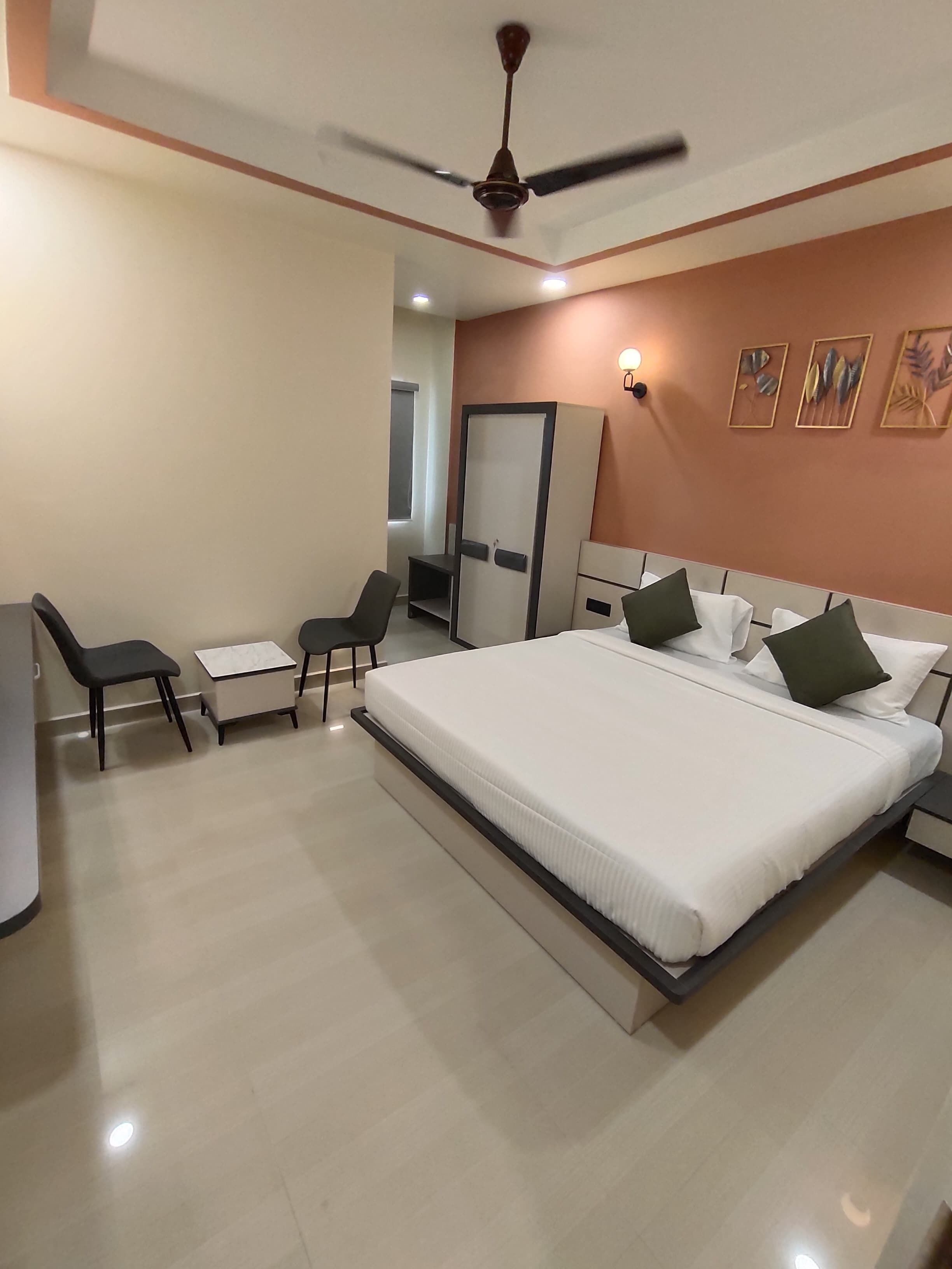 A modern hotel room with a large bed, a desk, two chairs, a wardrobe, and a ceiling fan.