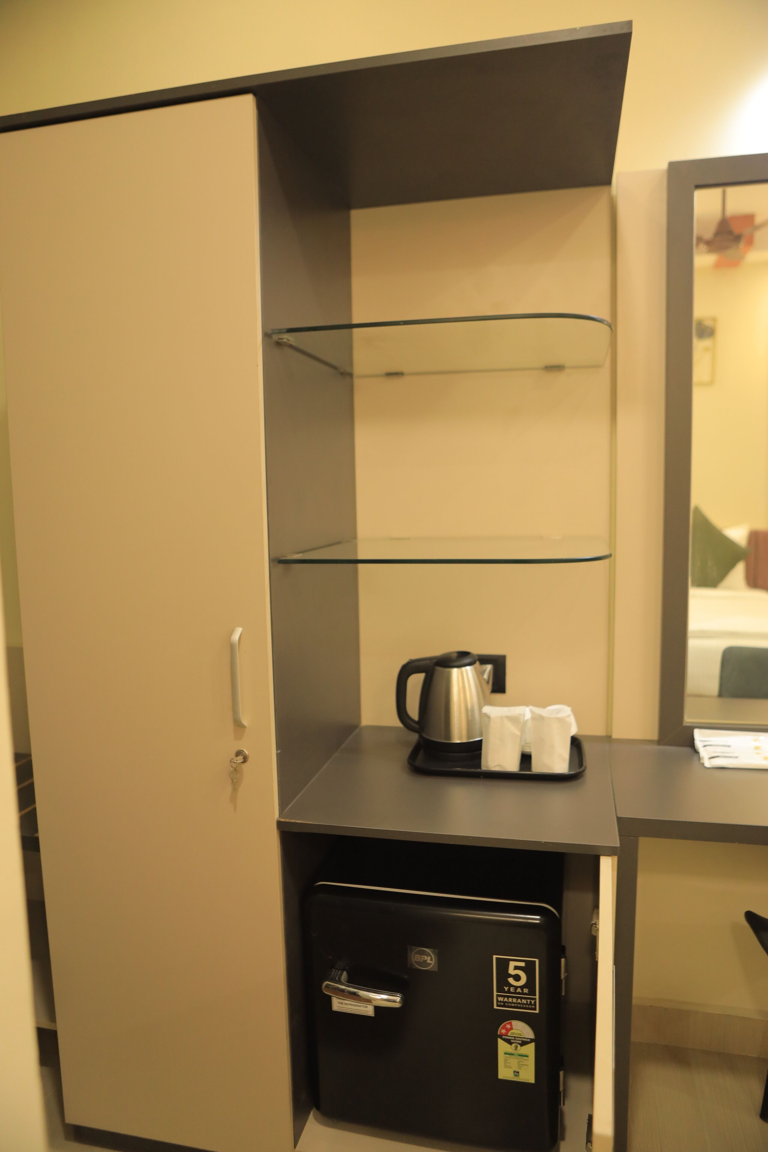 A compact hotel room with a minibar, a small safe, a glass shelf, and a mirror reflecting a bed.