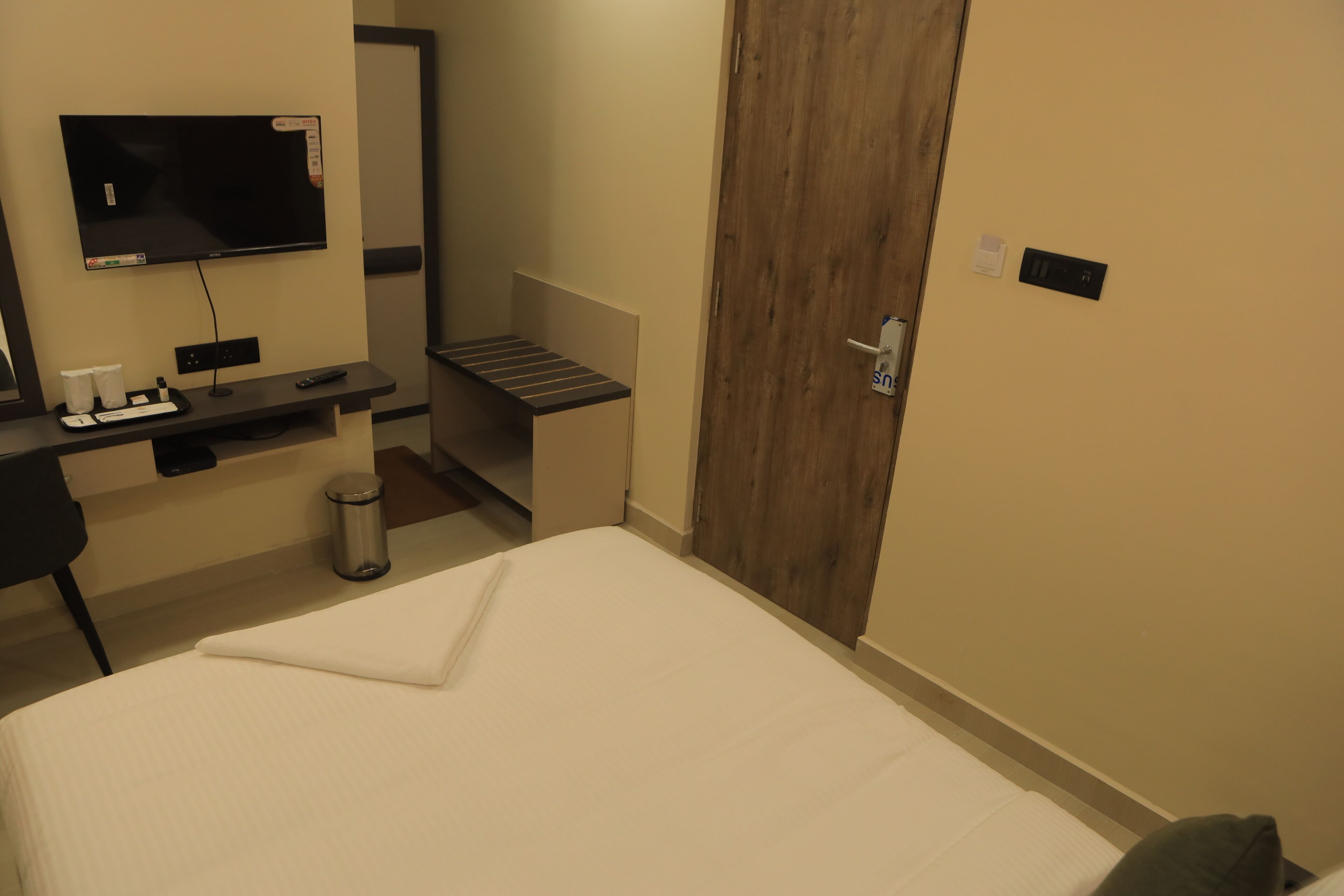 A hotel room with a bed, a desk with a flat-screen TV, a trash can, and a wooden door.