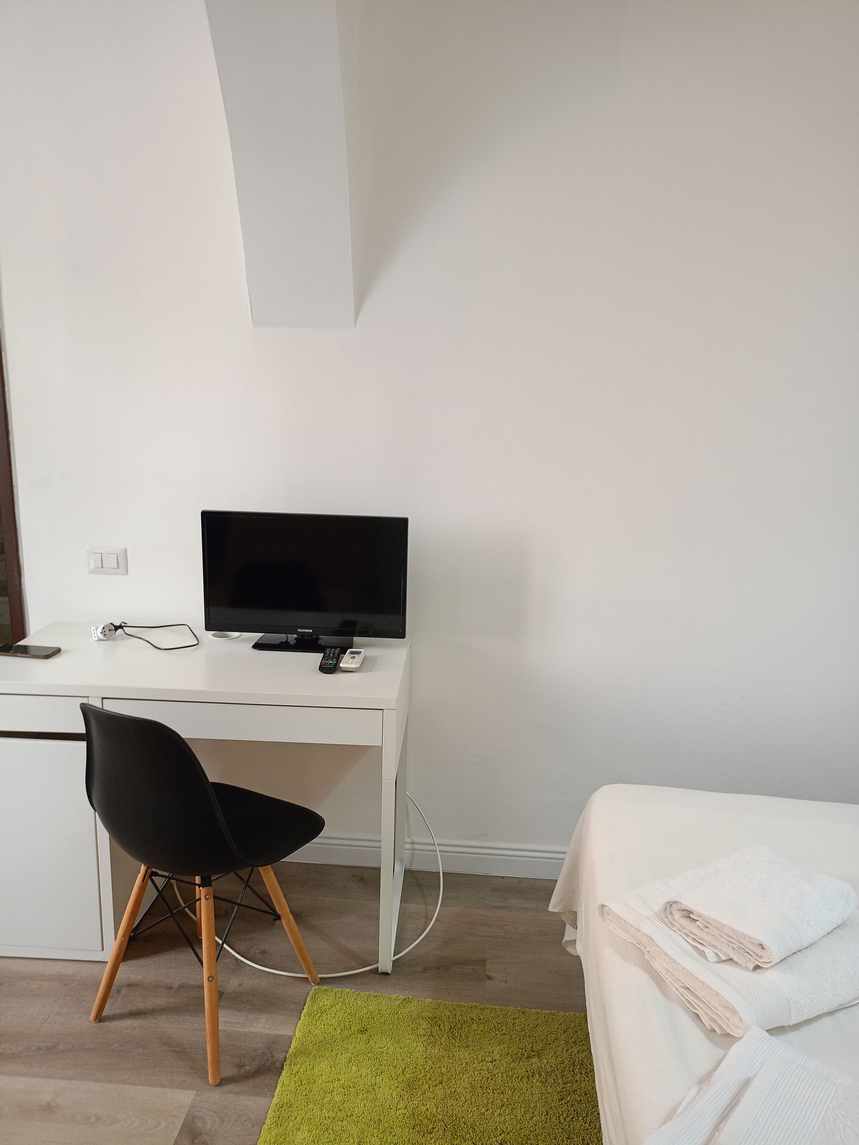A room with a white desk, a black chair, a bed with white bedding, and a green rug.