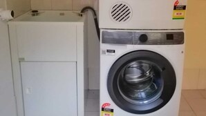 A compact laundry area with a stacked washer and dryer, a small shelf with baskets, and a tiled backsplash. - Adelaide Escape: Two-Storey Home Near City & Hills (Teringie)