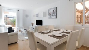 Un comedor moderno con una mesa blanca para cuatro personas, un televisor sobre un mueble blanco y un cuadro de un paisaje urbano en la pared.