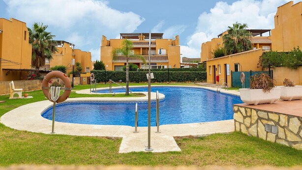 Una piscina rodeada de césped cuidado y palmeras, con un edificio con balcones al fondo.