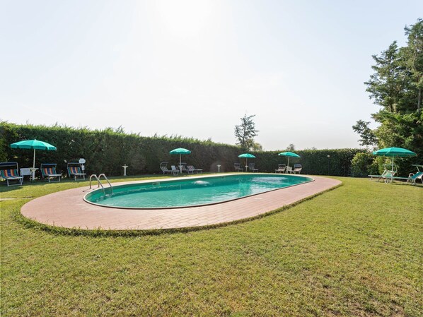 A rectangular swimming pool with a brick deck, surrounded by a grassy lawn and several lounge chairs with green umbrellas.