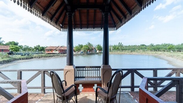 A covered outdoor seating area with two chairs and a table, overlooking a body of water and greenery. - Velankanni Lake Resort (Nagapattinam)