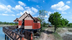 A small building with a red and beige exterior, a wooden bridge leading to it, and a tree nearby. - Velankanni Lake Resort (Nagapattinam)