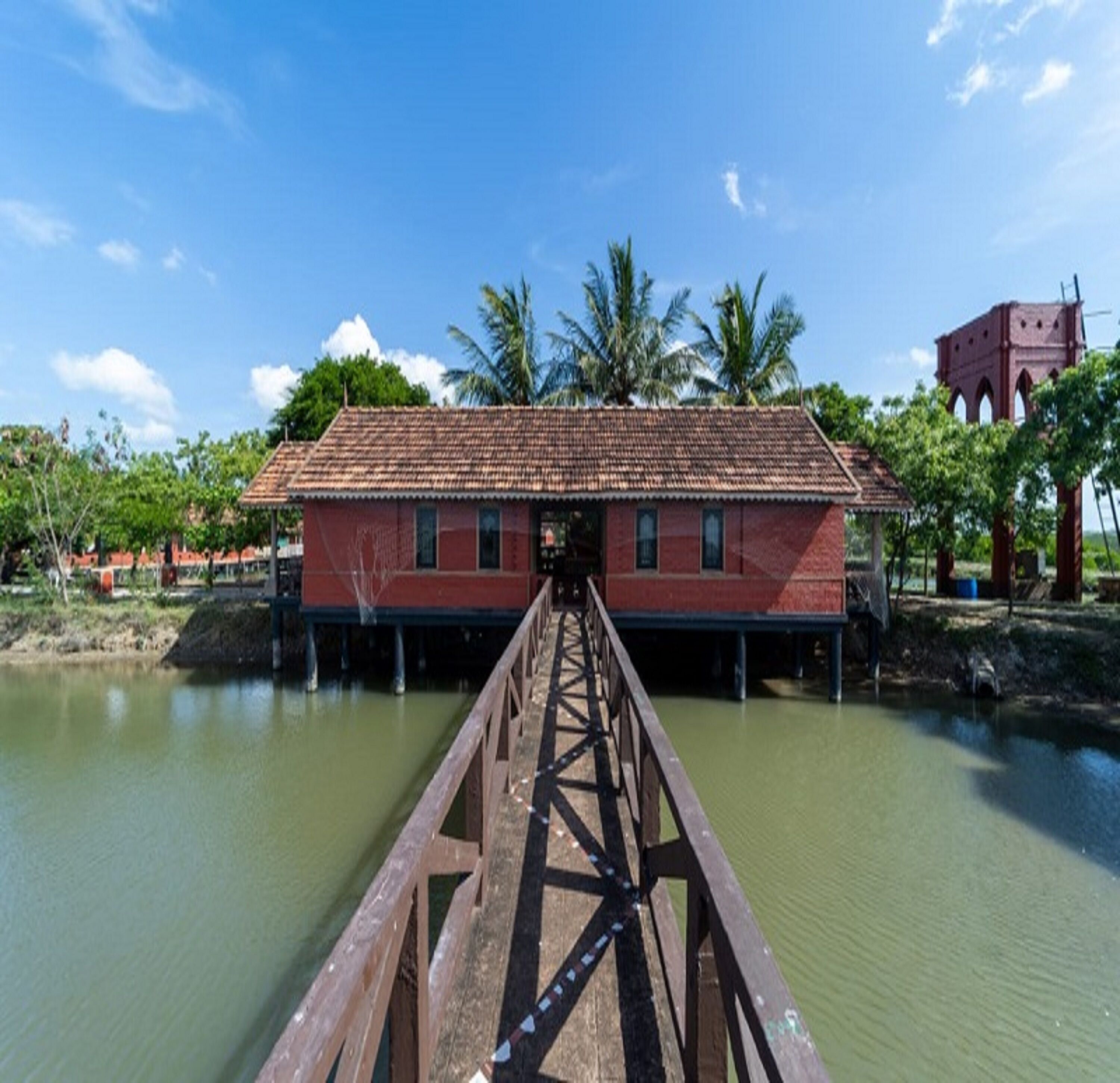 Un pont en bois mène à un bâtiment rouge doté d’un toit en tuiles, entouré d’eau et de palmiers.